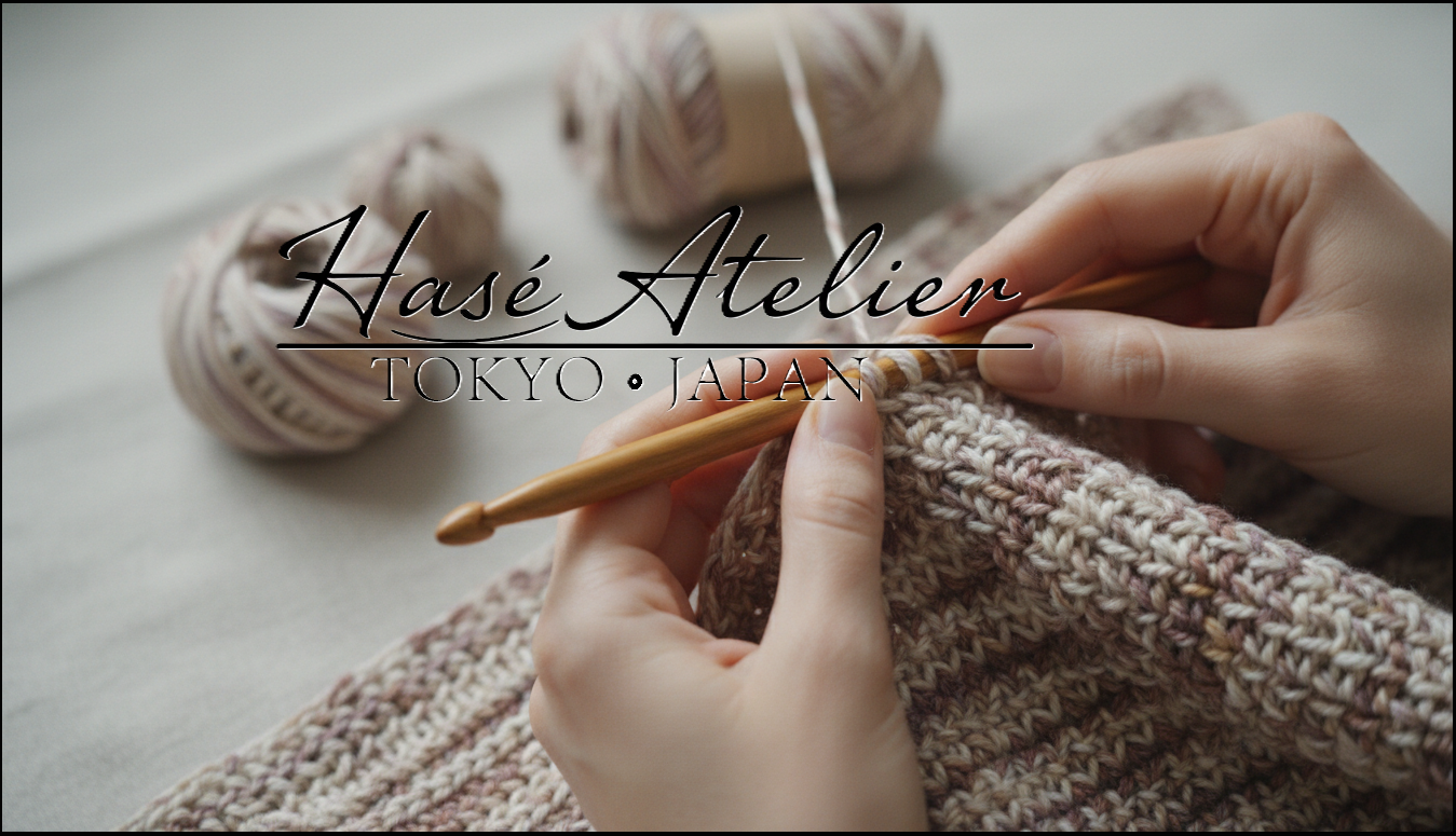 Close-up of hands crocheting with a wooden crochet hook and beige yarn, with a knitted fabric in soft neutral tones. The text 'Hasé Atelier TOKYO • JAPAN' is overlaid across the center.